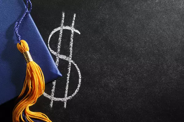 A blue graduation cap with a yellow tassel next to a dollar sign written in chalk