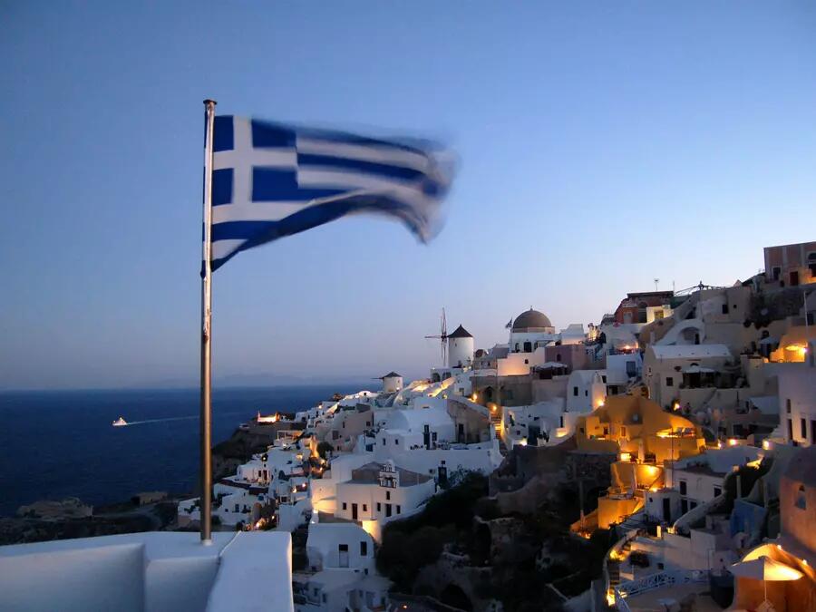 A Greek flag attached to a wall, with a hillside village and the sea in the background