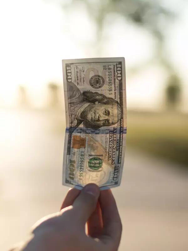 A hand holds up an American hundred-dollar bill, with a sunny background.