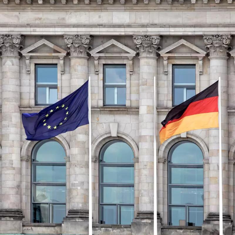 EU and German flags on flag poles in front of a stone building with large windows