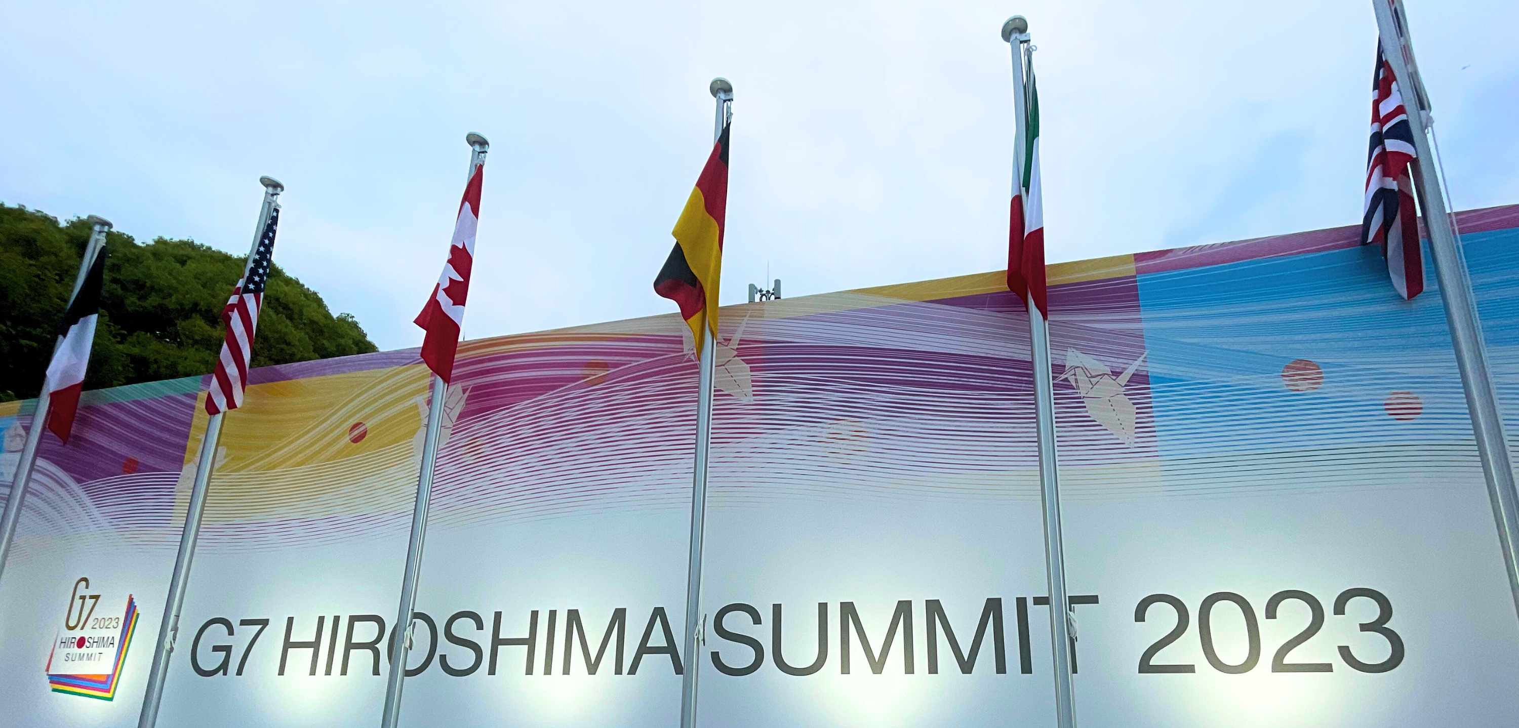 Flags of the G7 group are on tall flagpoles in front of a large display that says G 7 Summit Hiroshima 2023