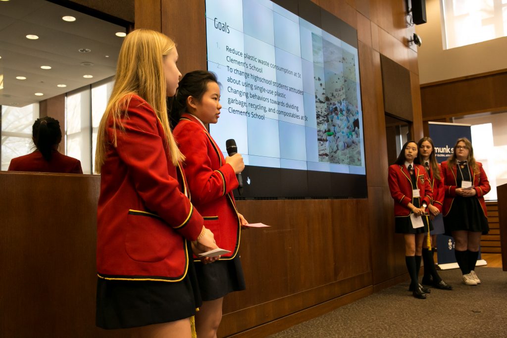 Five female students in school uniforms present in front of a large screen
