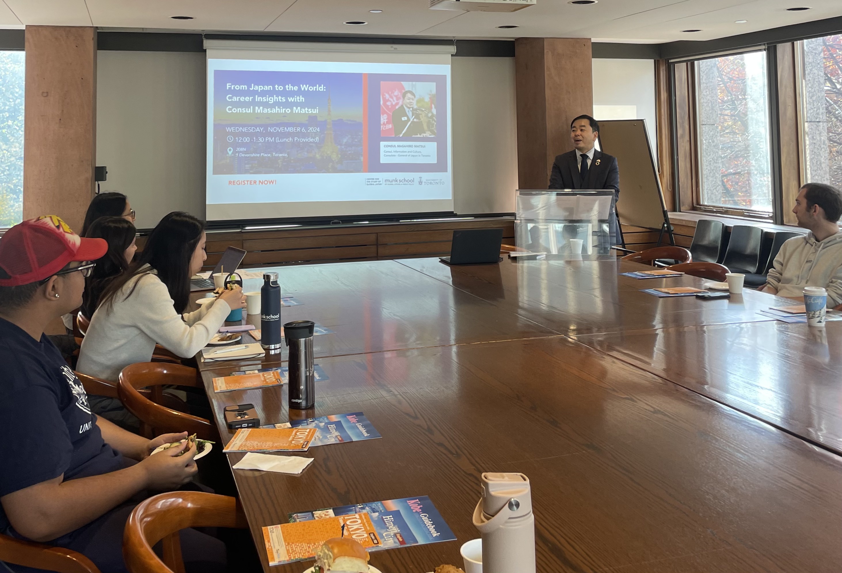 east asian man at podium giving talk to undergraduate students sat around table