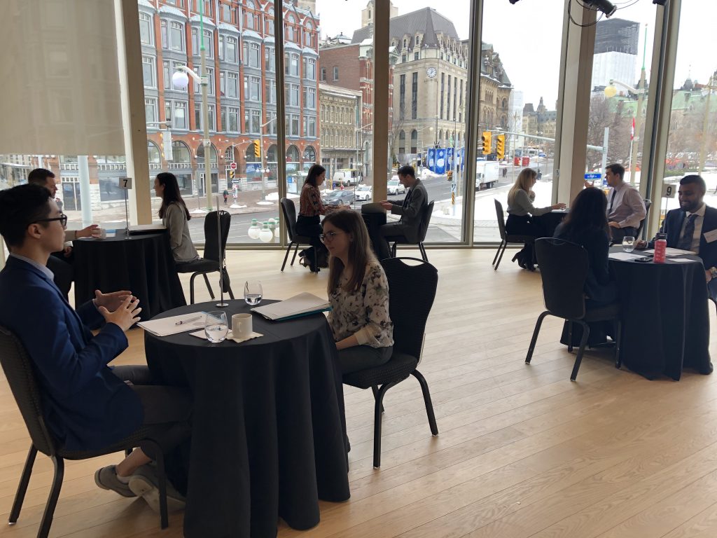 Students talk in pairs at round tables with large windows and Ottawa streetscape in the background