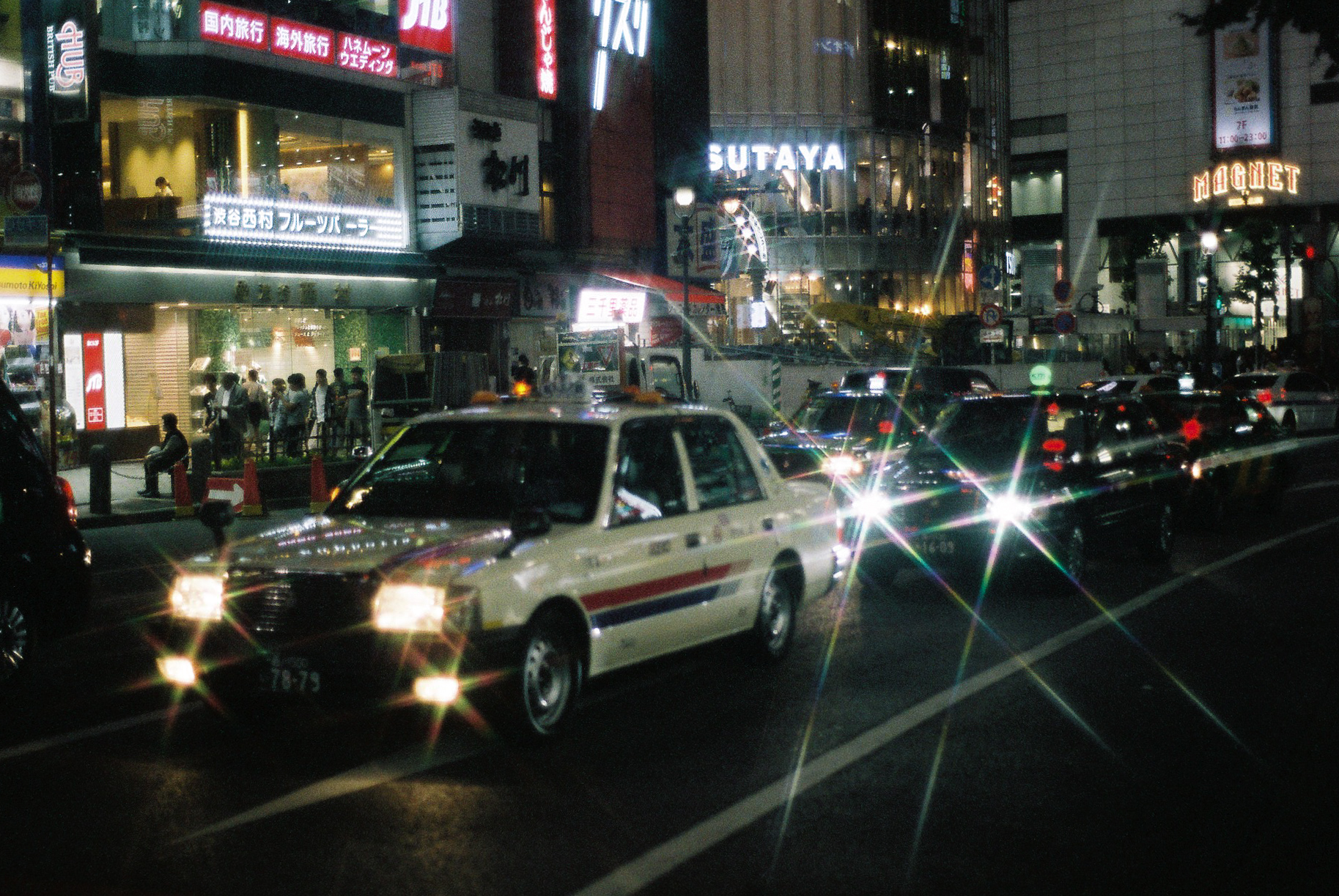 A photo of a street in Tokyo at night time capturing a taxi driving by with its bright headlights on.