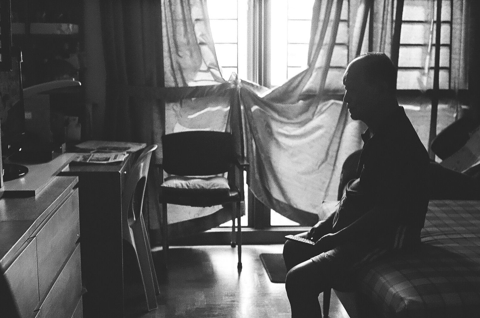 Black and white photo of an elderly man seating still on the edge of a bed