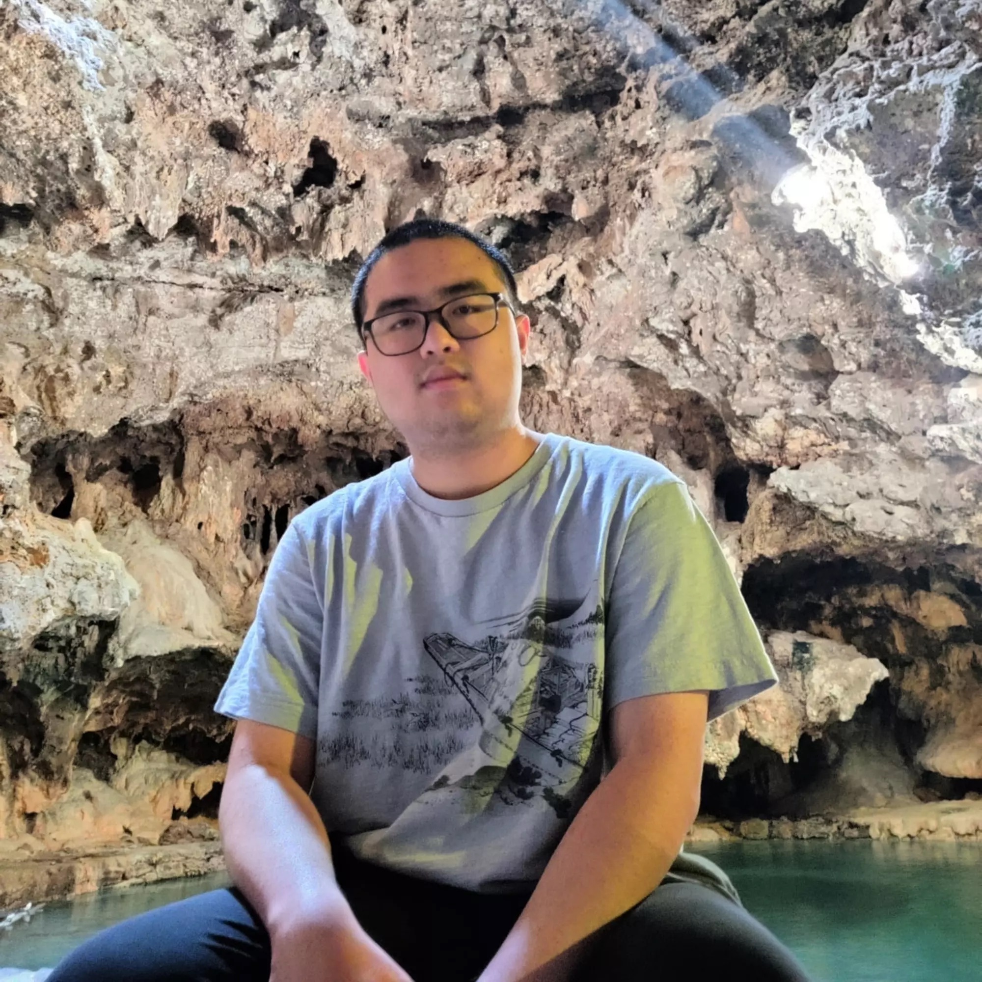 East Asian man sitting in front of rocks wearing grey T-shirt