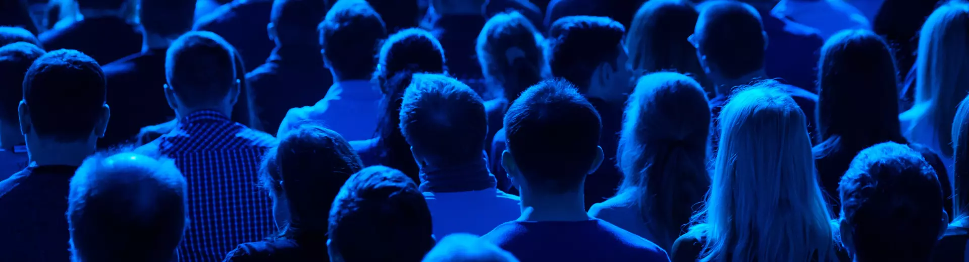Photo of the back of heads of a crowd of people sitting in rows of chairs in a blue light 