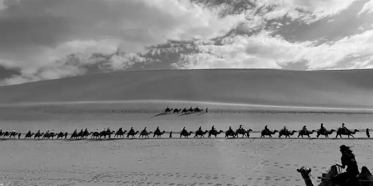 Black and white landscape image of a sand dune with clouds in the sky above. A line of people riding camels appears across the horizon, with one person on a camel in the foreground.
