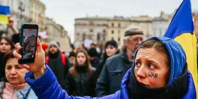 A women wearing a blue jacket with a hood and "stop war" across her cheeks holds up a phone to record the anti-war protest