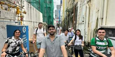 CAS students on bicycles in a narrow alleyway in Singapore's Chinatown, surrounded by historic buildings. 