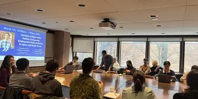 Picture of a lecture with students around a table and a presenter. 