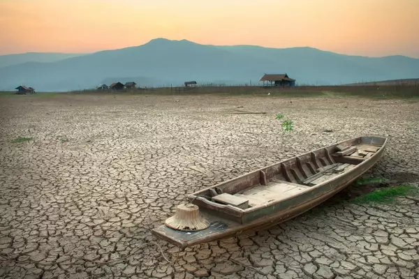 A worn row boat sits on parched, cracked land with mountains and a sunset in the background