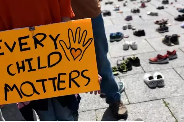 Child holding "every child matters" sign with shoes in the background on the cement