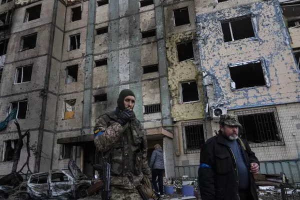 Two Ukrainian men, one in full military garb and one wearing plain clothes with an armband and military cap smoke cigarettes infront of a bombed out building