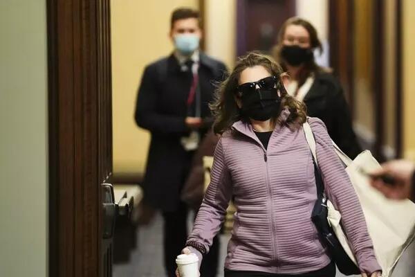 Chrystia Freeland wearing a black mask, sunglasses, a light purple zip up sweater with a tote bag and coffee cup in hand is flanked by two masked aides, presumably walking inside the Parliament building