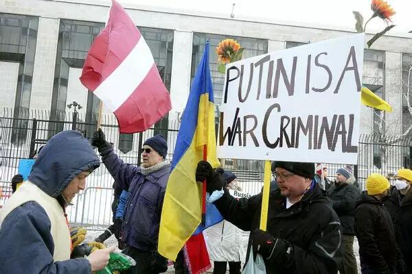 Protestors hold various flags, including a Ukrainian flag and hold a large sign that says "Putin is a War Criminal"