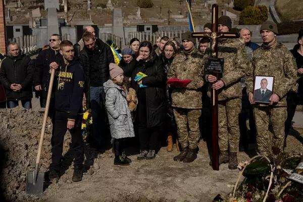 Ukrainians participating in a burial. Some are wearing military clothes, and one is holding a large cross, another holding a photograph of a uniformed man, presumed to be deceased.