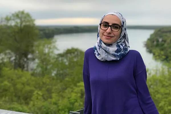Photo of Noura Al-Jizawi posed in front of a body of water and trees from the waist up. She wears a white and blue patterned hijab and a dark blue long sleeve shirt. She has a soft smile and is wearing square-shaped glasses.