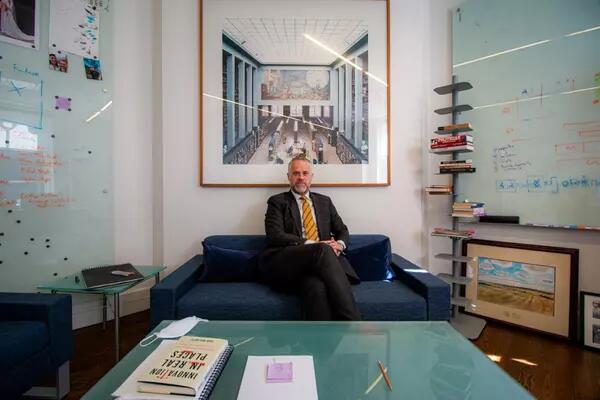 Peter Loewen sits crossed-legged in an office. He is wearing a suit with a tie and is flanked by a book shelf on the right and a glass surface with writing.