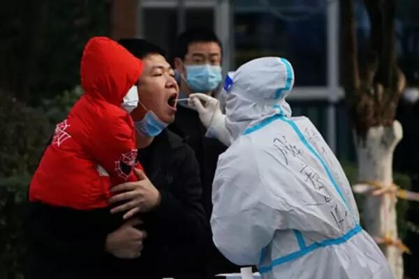 A man holding a masked child gets swabbed for COVID by a worker in a full sanitation suit