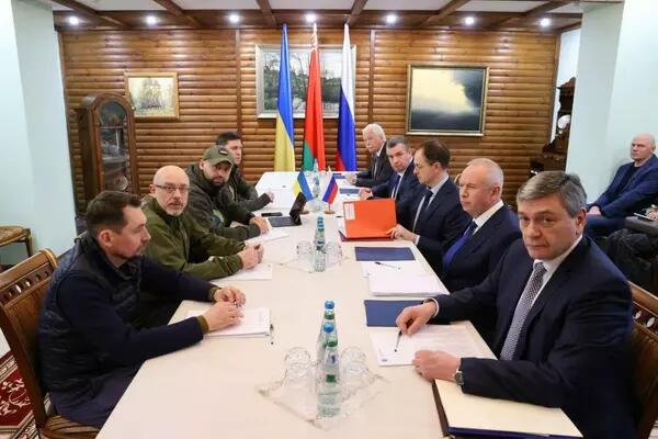 Politicians sit around a long table in negotiation, with the Ukrainian, Russian, and a red and green flag in the background