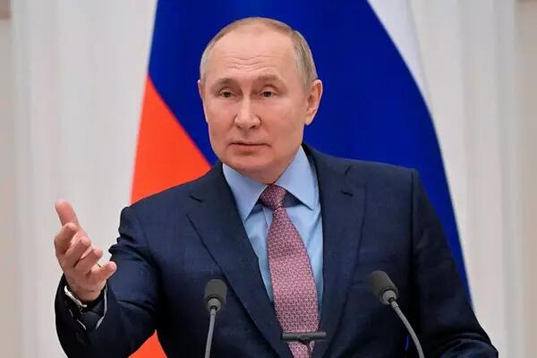 Vladimir Putin infront of a Russian flag speaking to a crowd out of sight, wearing a navy blue suit, light blue shirt and red patterned tie