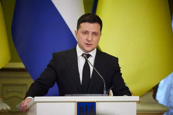 President Volodymyr Zelensky wears a black suit and tie and is speaking at a podium with flags behind him.