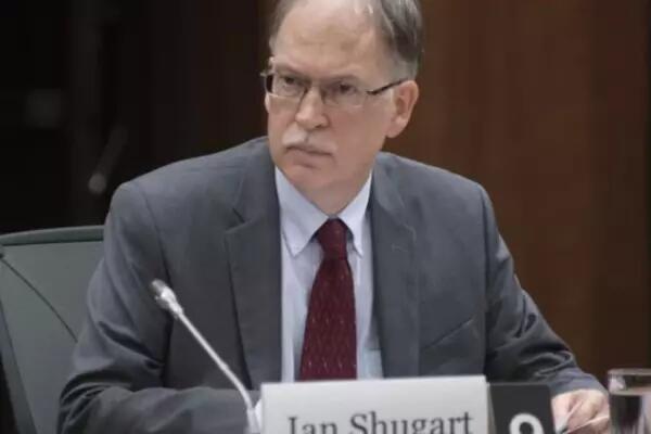 Ian Shugart wears a gray suit with a burgundy tie and square eyeglasses