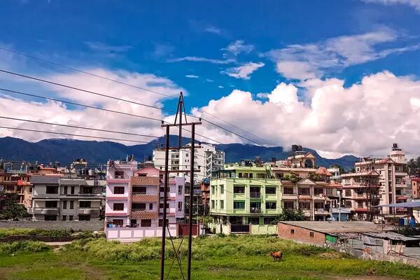 A green field and telephone pole foregrounds a town on colourful pink and green low-rise buildings in Nepal.