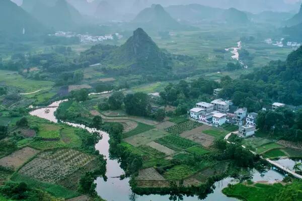 A blue river winds through the green countryside of Guilin, China while pointed hills rise in the distance against the pink and purple sunset.