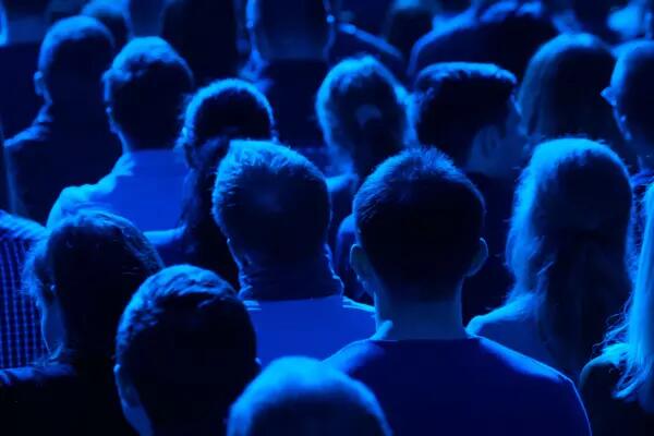 Photo of the back of heads of a crowd of people sitting in rows of chairs in a blue light 