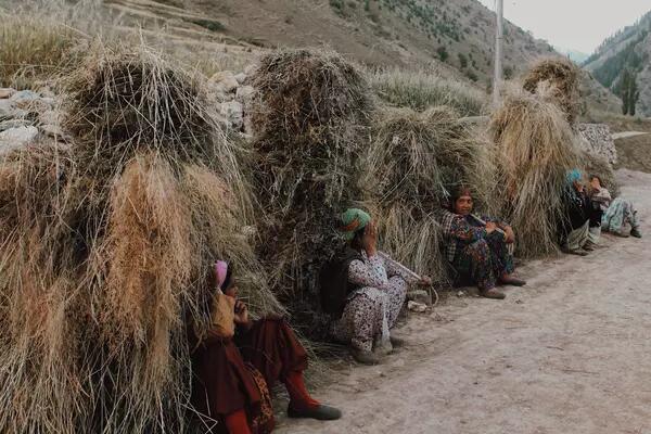 Women sit by the side of a dirt road with piles of hay behind their backs.