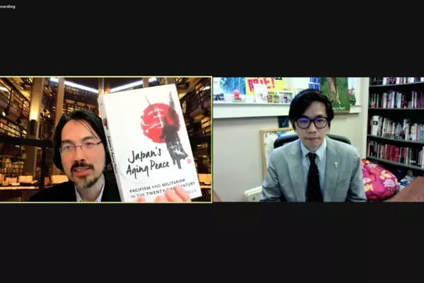 A screenshot of two panelists having a discussion, the one on the left is holding up a book