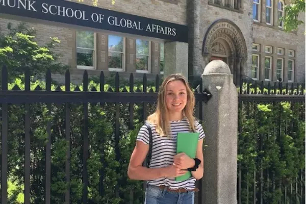 Picture of Abigail McDonald standing in front of the Munk School of Global Affairs, wearing blue jeans and a stripped t-shirt.