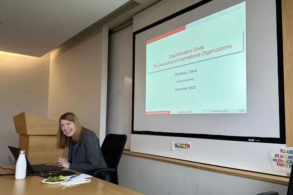 Woman sitting at a table smiling in front of a powerpoint presentation.