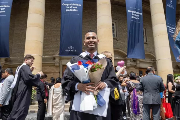 Jaivet Ealom stand in front of convocation hall in his graduation regalia holding flowers and his diploma