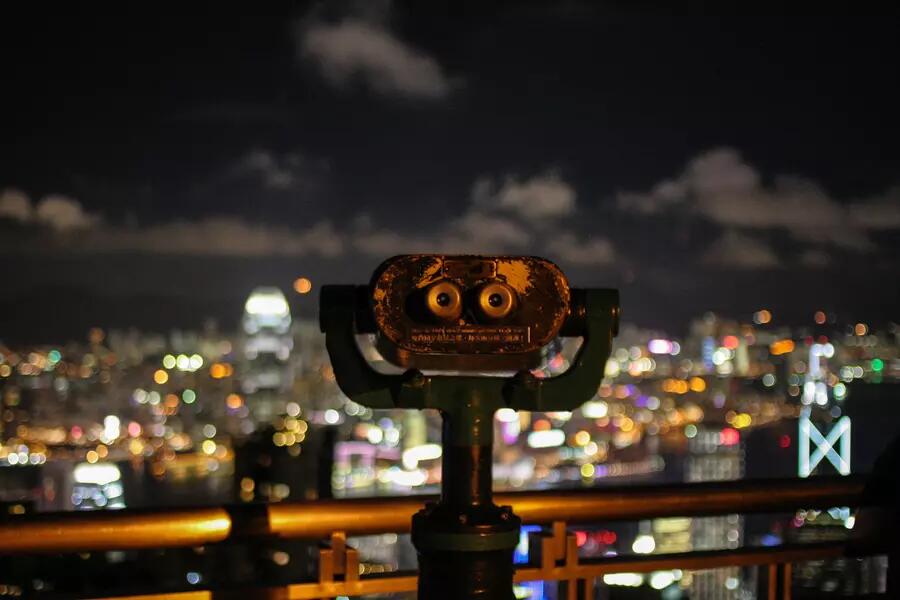 A pair of binoculars looks out on a lit up city below at night. 