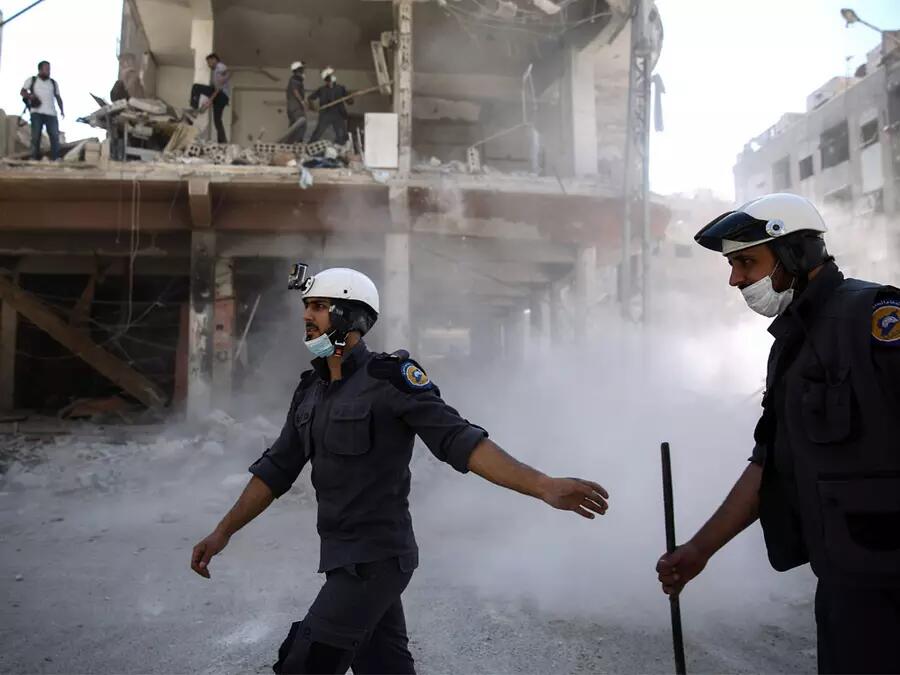 Two members of the White Helmets walk past smoke and a collapsed building