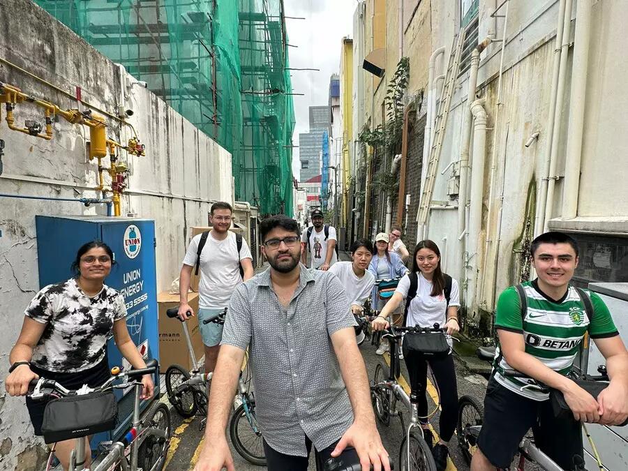 CAS students on bicycles in a narrow alleyway in Singapore's Chinatown, surrounded by historic buildings. 