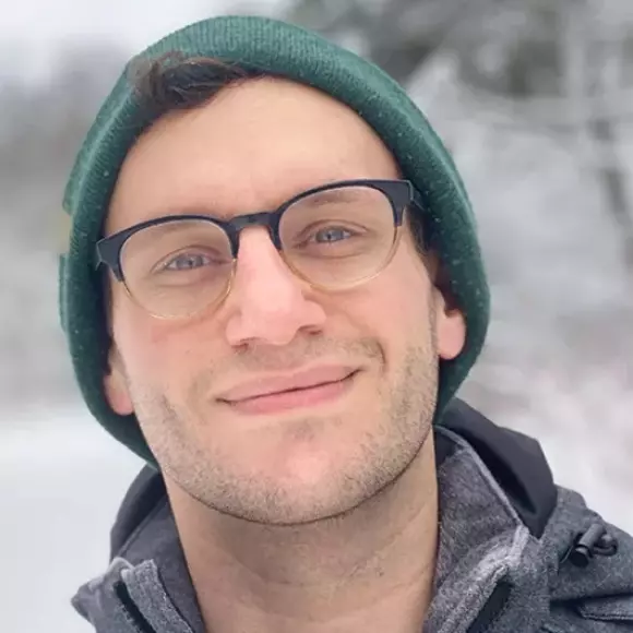 Picture of Dan Guadagnolo wearing a grey winter jacket, glasses, and a green hat with a snowy winter background.