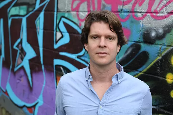 Picture of Kevin O'Neill standing in front of a graffiti wall and wearing a blue collar shirt. 