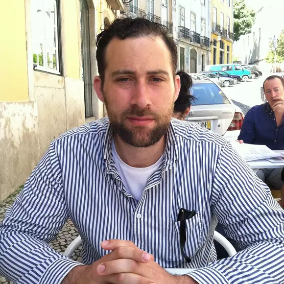Picture of Max Mishler sitting at a table outdoors and wearing a blue and white stripped shirt. 