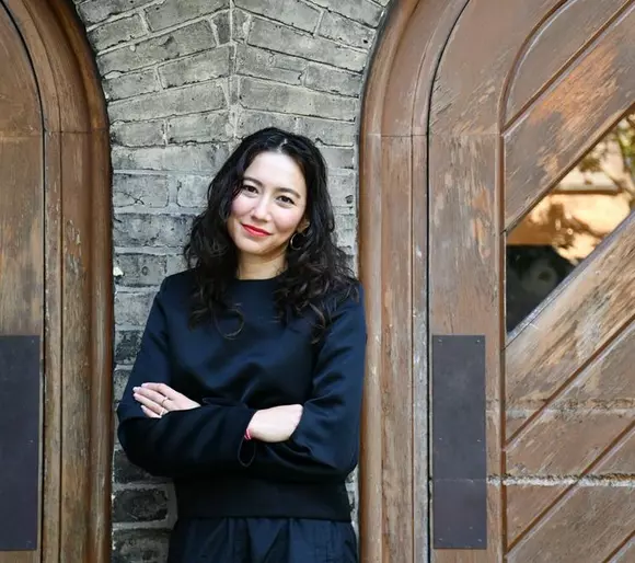 Picture of Maya Harakawa wearing a black long sleeve shirt with a grey brick background.