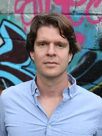 Picture of Kevin O'Neill standing in front of a graffiti wall and wearing a blue collar shirt. 