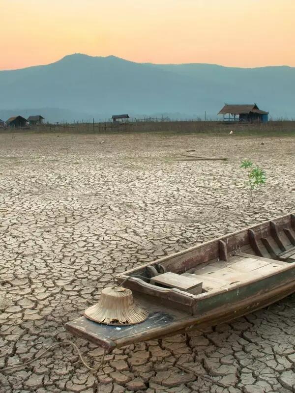 A worn row boat sits on parched, cracked land with mountains and a sunset in the background