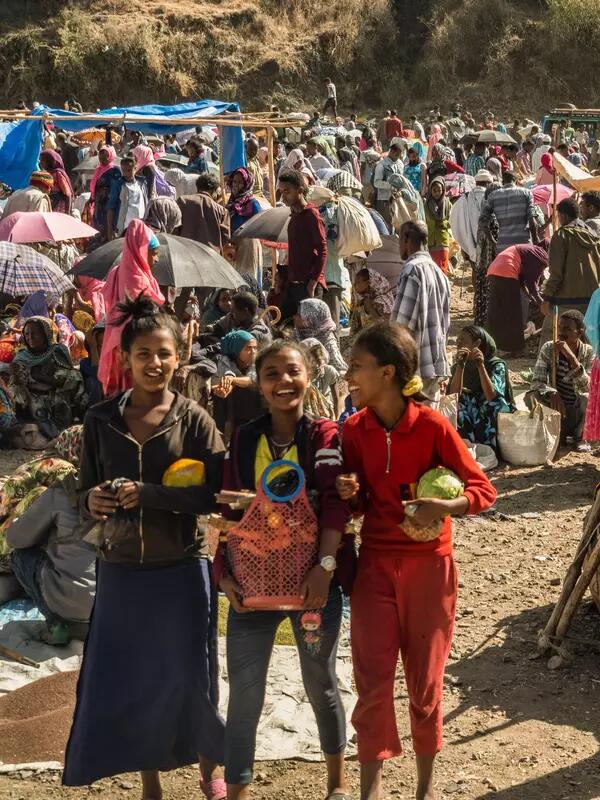 Three laughing girls holding food baskets in Ethiopia, against the backdrop of a large crowd