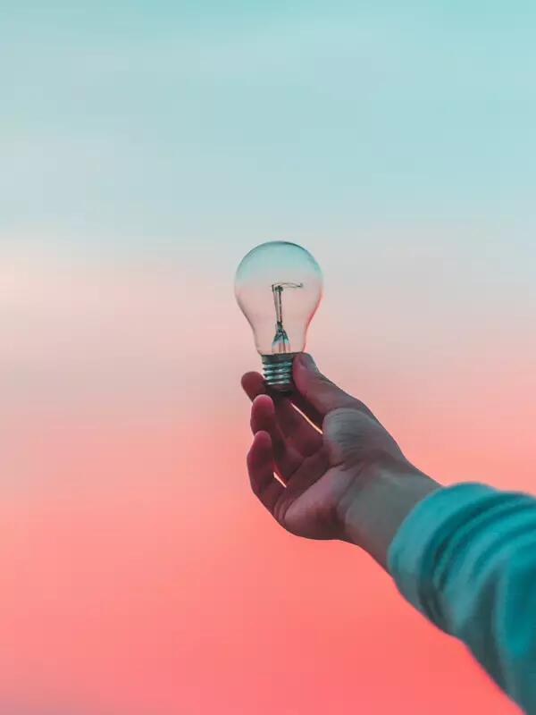 A hand holds a lightbulb up against a pink and blue sunset.