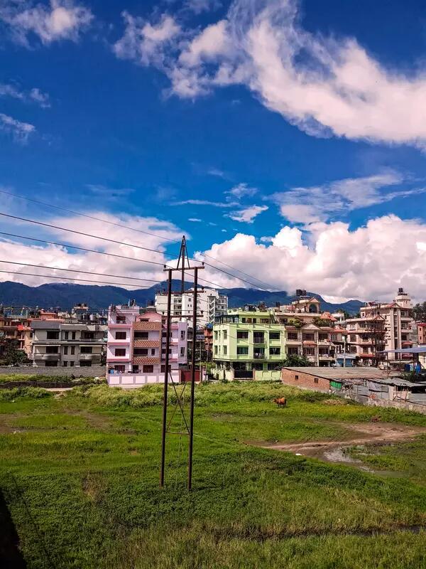 A green field and telephone pole foregrounds a town on colourful pink and green low-rise buildings in Nepal.