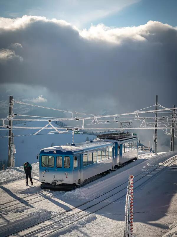A blue and white two-car train sits on snowy tracks. A man walks in front and cables hang overhead. The sky is cloudy.